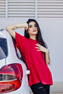 A person with long dark hair wearing a bright red shirt leans against a white car. The background consists of a neutral wall with horizontal lines.