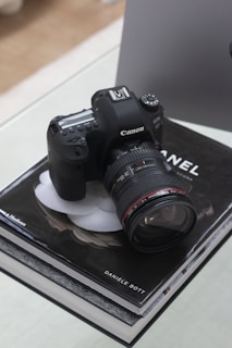 A black Canon EOS 6D camera is resting on top of a Chanel book by Danièle Bott. The camera is equipped with a large lens and placed on a glass surface. There is a blurred background with neutral tones.