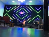 A modern interior setup with vibrant, geometric neon lights in green and blue illuminating the background. The center stage has a small screen displaying colorful graphics, alongside a lit podium with floral decorations. Musical instruments like drums are visible on one side, with various chairs arranged in the foreground.