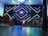 A modern interior setup with vibrant, geometric neon lights in green and blue illuminating the background. The center stage has a small screen displaying colorful graphics, alongside a lit podium with floral decorations. Musical instruments like drums are visible on one side, with various chairs arranged in the foreground.