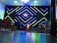 A modern interior setup with vibrant, geometric neon lights in green and blue illuminating the background. The center stage has a small screen displaying colorful graphics, alongside a lit podium with floral decorations. Musical instruments like drums are visible on one side, with various chairs arranged in the foreground.