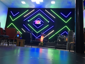 A modern interior setup with vibrant, geometric neon lights in green and blue illuminating the background. The center stage has a small screen displaying colorful graphics, alongside a lit podium with floral decorations. Musical instruments like drums are visible on one side, with various chairs arranged in the foreground.