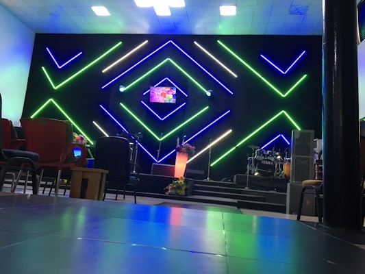 A modern interior setup with vibrant, geometric neon lights in green and blue illuminating the background. The center stage has a small screen displaying colorful graphics, alongside a lit podium with floral decorations. Musical instruments like drums are visible on one side, with various chairs arranged in the foreground.