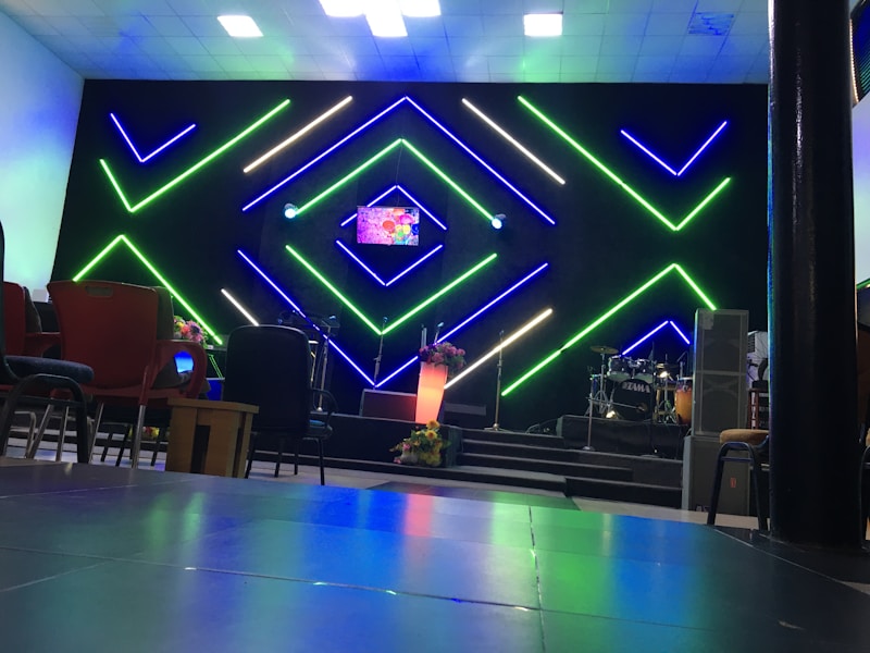 A modern interior setup with vibrant, geometric neon lights in green and blue illuminating the background. The center stage has a small screen displaying colorful graphics, alongside a lit podium with floral decorations. Musical instruments like drums are visible on one side, with various chairs arranged in the foreground.