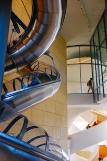 A modern architectural space featuring a large, shiny metal slide winding through an indoor atrium. The interior is composed of glass walls and doors, along with smooth, cream-colored walls and ceilings. People are seen walking around and ascending stairs, which have bright orange steps. The overall design combines contemporary and industrial elements.