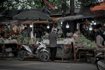 A busy outdoor market stall scene with several people engaged in activities around the stall. The stall is covered with a canopy and is selling various fresh vegetables and herbs. There are plastic bags filled with produce, hanging items, and various fresh greens displayed. A motorcycle is parked in front of the stall, and there are hanging lights illuminating the area.