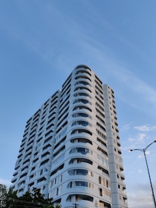 A modern, high-rise residential building with a sleek design featuring rounded balconies and a white facade. The structure stands tall against a clear blue sky with slight wisps of clouds. Streetlights and some greenery are visible at the base of the building.
