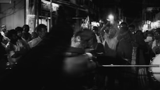 A black and white image of a group of people gathered on a street at night. They appear to be participating in a procession or festival, with some individuals playing musical instruments and others carrying objects or taking photos. The scene is lively and crowded, with a sense of movement and energy.