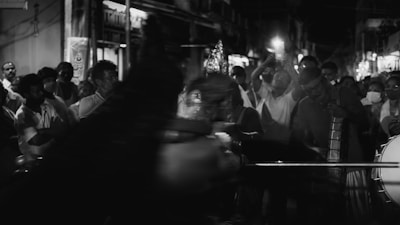 A black and white image of a group of people gathered on a street at night. They appear to be participating in a procession or festival, with some individuals playing musical instruments and others carrying objects or taking photos. The scene is lively and crowded, with a sense of movement and energy.