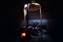 Inside a dimly lit yurt, a person sits on a bench, appearing contemplative. The space is warmly illuminated by a small stove, casting a cozy glow. The interior features wooden poles and a circular skylight that provides natural light.