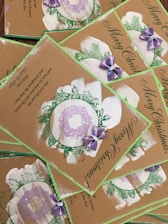 A collection of Christmas cards with a rustic design, featuring green foliage, purple bows, and wreath accents. The cards bear the message 'Merry Christmas' in elegant script, and the background is a natural brown, giving them a hand-crafted appearance.