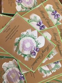 A collection of Christmas cards with a rustic design, featuring green foliage, purple bows, and wreath accents. The cards bear the message 'Merry Christmas' in elegant script, and the background is a natural brown, giving them a hand-crafted appearance.