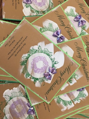 A collection of Christmas cards with a rustic design, featuring green foliage, purple bows, and wreath accents. The cards bear the message 'Merry Christmas' in elegant script, and the background is a natural brown, giving them a hand-crafted appearance.