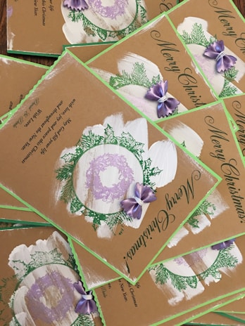 A collection of Christmas cards with a rustic design, featuring green foliage, purple bows, and wreath accents. The cards bear the message 'Merry Christmas' in elegant script, and the background is a natural brown, giving them a hand-crafted appearance.