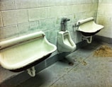 A public restroom with two wall-mounted urinals and a sink on a concrete floor. The walls are made of light-colored stone tiles, and there are visible stains and discoloration on the floor.