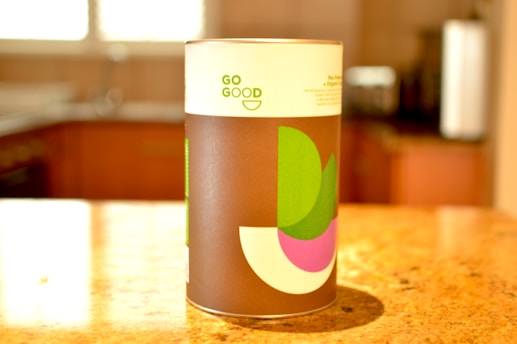 A cylindrical container with a modern design label on a kitchen countertop, featuring geometric shapes in green, purple, and white against a brown background. The words 'GO GOOD' are printed on the label.