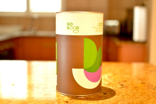 A cylindrical container with a modern design label on a kitchen countertop, featuring geometric shapes in green, purple, and white against a brown background. The words 'GO GOOD' are printed on the label.