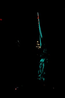 A predominantly dark image featuring a tall, illuminated structure with teal and red lighting. Smaller lights and subtle details are visible around the main structure, adding to the overall ethereal and mysterious atmosphere.