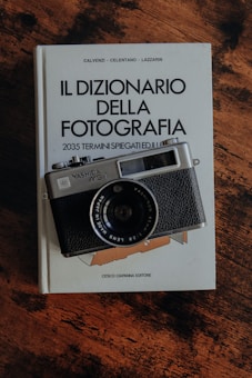 A vintage Yashica MG-1 camera rests on an open book titled 'Il Dizionario della Fotografia'. The book has a white cover with text and is lying on a wooden surface. The lighting creates a warm, nostalgic ambiance.