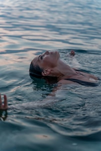 A woman is floating in a body of water, with her eyes closed and face relaxed, suggesting a sense of peace and tranquility. The water is calm and reflects the natural light, creating a serene and soothing atmosphere.