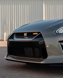 The front section of a sleek, modern sports car with an aggressive design. Prominent brand logos and detailed grille features are visible. The background includes a vertical lined wall, enhancing the car's sharp and dynamic appearance.