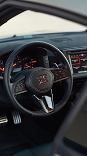 A close-up view of a car's interior focusing on the steering wheel, which has a distinctive logo in the center. The dashboard displays multiple gauges, and there is a digital screen on the right side showing infotainment controls. The pedals can be seen at the bottom, and the overall design appears sleek and modern.