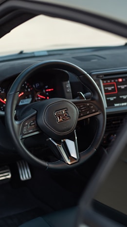 A close-up view of a car's interior focusing on the steering wheel, which has a distinctive logo in the center. The dashboard displays multiple gauges, and there is a digital screen on the right side showing infotainment controls. The pedals can be seen at the bottom, and the overall design appears sleek and modern.