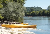 Several yellow canoes are resting on a rocky riverbank under the shade of a large tree. The river flows smoothly with groups of people kayaking in the distance. Lush, green trees line the opposite bank against a backdrop of gentle hills under a clear blue sky.