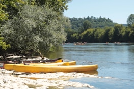 Several yellow canoes are resting on a rocky riverbank under the shade of a large tree. The river flows smoothly with groups of people kayaking in the distance. Lush, green trees line the opposite bank against a backdrop of gentle hills under a clear blue sky.