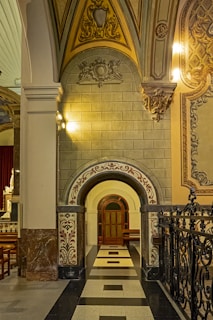 An ornate architectural interior with intricate designs and ornate carvings. The image features a stone archway with detailed patterns and decorative moldings on the ceiling and walls. Warm lighting highlights the textures and carvings, creating a sense of grandeur.