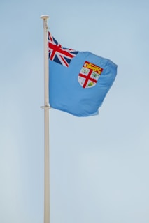 A light blue flag with the Union Jack in the top left corner and a shield emblem featuring a lion and plants on the right side, attached to a flagpole against a clear sky.