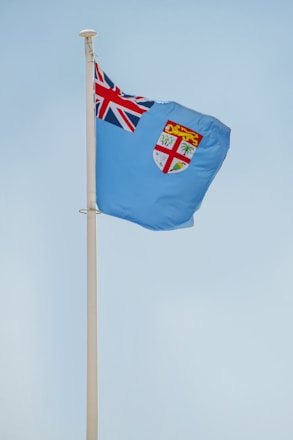 A light blue flag with the Union Jack in the top left corner and a shield emblem featuring a lion and plants on the right side, attached to a flagpole against a clear sky.