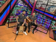 Two individuals are seated in a vibrant, colorful setting, with one holding a guitar and the other sitting on a cajón drum. The background features dynamic, abstract artwork with various colors and patterns. The individuals are casually dressed, suggesting a musical performance or rehearsal.