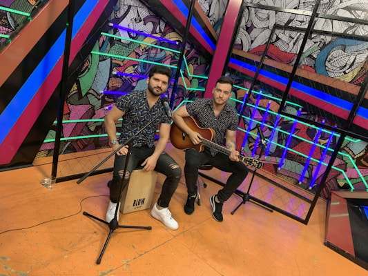 Two individuals are seated in a vibrant, colorful setting, with one holding a guitar and the other sitting on a cajón drum. The background features dynamic, abstract artwork with various colors and patterns. The individuals are casually dressed, suggesting a musical performance or rehearsal.
