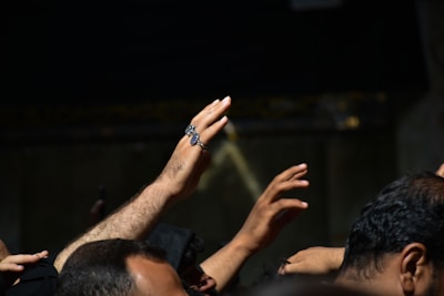 Several hands are raised against a dark background, with focus on one hand adorned with a distinctive ring.
