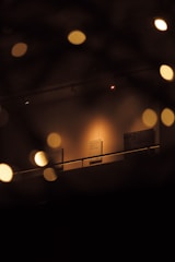 An art gallery or museum space dimly lit with warm lights. Several artworks or informational placards hang on the walls. The view is slightly obscured by out-of-focus circular lights, creating a bokeh effect.