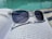 A pair of transparent sunglasses with dark lenses rests on an open book with printed text, positioned near a swimming pool. The sunlight creates reflections on the water and the surface of the book.