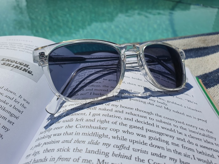 A pair of transparent sunglasses with dark lenses rests on an open book with printed text, positioned near a swimming pool. The sunlight creates reflections on the water and the surface of the book.