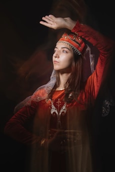 A person dressed in traditional attire with intricate embroidery and a decorative headpiece. The warm lighting highlights the rich red and gold colors of the clothing, while the dark background adds depth. The pose is graceful and the person appears to be looking upwards with a contemplative expression.