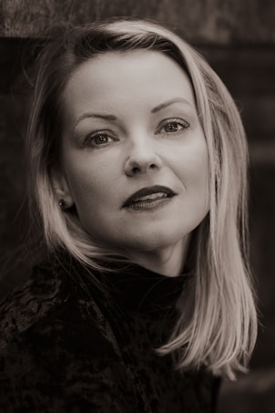 A black and white portrait of a woman with light hair and subtle expression, wearing a dark textured top. Her gaze is directed slightly above the camera and she has an engaged, thoughtful look.