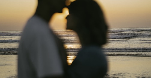 A silhouetted couple stands close together in front of a sunset with the ocean waves in the background. The warm tones of the sunset create a romantic and serene atmosphere.
