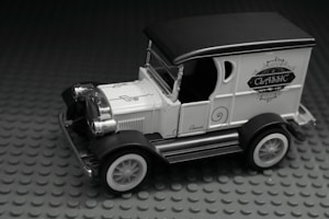 A miniature vintage car model with ornate detailing, featuring a black roof, white body, and classic design elements. The car is displayed on a textured gray base.