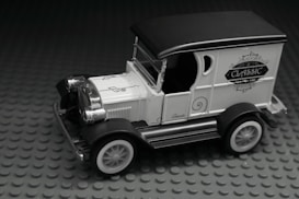 A miniature vintage car model with ornate detailing, featuring a black roof, white body, and classic design elements. The car is displayed on a textured gray base.