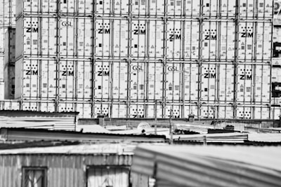 Large stacks of shipping containers, branded with 'ZIM', are tightly packed together, creating a grid-like pattern. In the foreground, the tops of corrugated metal roofs from nearby structures are visible.