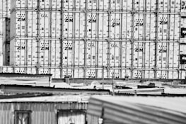 Large stacks of shipping containers, branded with 'ZIM', are tightly packed together, creating a grid-like pattern. In the foreground, the tops of corrugated metal roofs from nearby structures are visible.