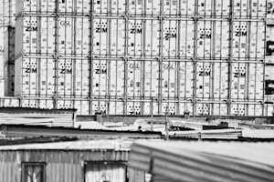 Large stacks of shipping containers, branded with 'ZIM', are tightly packed together, creating a grid-like pattern. In the foreground, the tops of corrugated metal roofs from nearby structures are visible.
