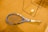 A tennis racket with a blue frame lies on a brown, textured surface, alongside a tennis ball. There are metal frame structures casting shadows across the scene, suggesting an indoor setting.