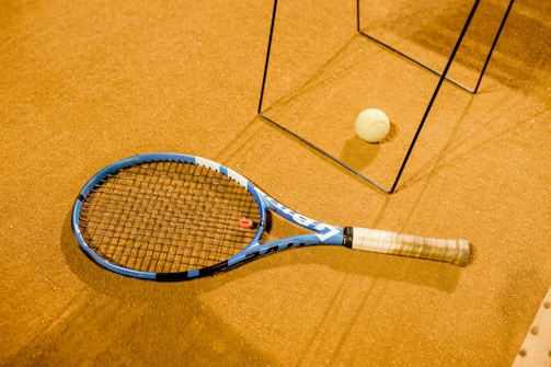 A tennis racket with a blue frame lies on a brown, textured surface, alongside a tennis ball. There are metal frame structures casting shadows across the scene, suggesting an indoor setting.