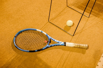 A tennis racket with a blue frame lies on a brown, textured surface, alongside a tennis ball. There are metal frame structures casting shadows across the scene, suggesting an indoor setting.