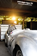 A vintage 1954 Cooper-Jaguar T33 car parked under a canopy with a warm sunset filtering through trees in the background. The car, with the number 34 displayed on its side, reflects the golden light, accentuating its sleek silver body. Tools and equipment are seen on a cabinet behind the car, suggesting a setting for maintenance or racing.